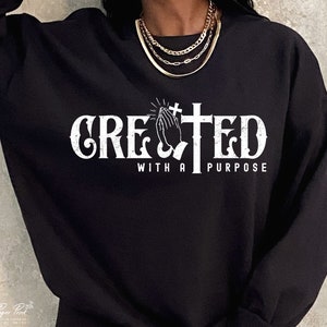 May include: Black sweatshirt with a white graphic design that says "CREATED WITH A PURPOSE". The graphic features a cross and praying hands.