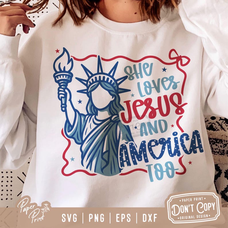 Puede incluir: Una sudadera blanca con un dise&ntilde;o gr&aacute;fico rojo, blanco y azul. El gr&aacute;fico presenta a una mujer con un disfraz de la Estatua de la Libertad, una antorcha y el texto "She Loves Jesus and America Too".