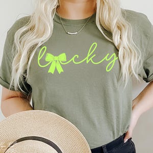 Puede incluir: Una camiseta verde con la palabra "lucky" escrita en verde ne&oacute;n con un lazo verde. La modelo lleva la camiseta y sostiene un sombrero de paja.