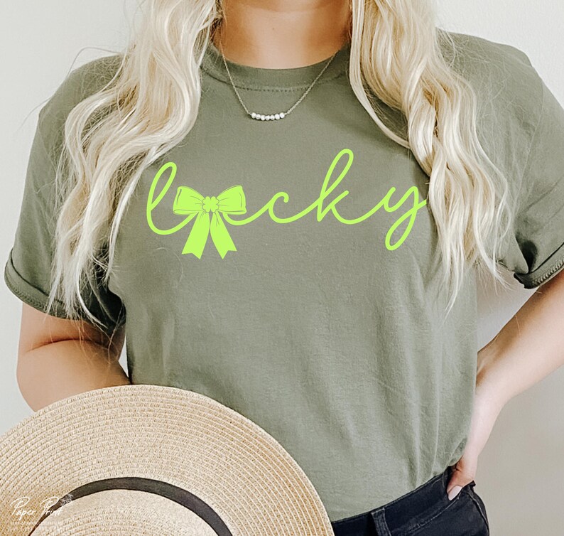 Puede incluir: Una camiseta verde con la palabra "lucky" escrita en verde ne&oacute;n con un lazo verde. La modelo lleva la camiseta y sostiene un sombrero de paja.