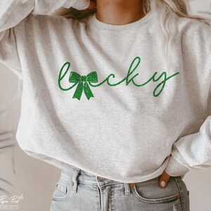 Puede incluir: Una sudadera gris con un dise&ntilde;o de purpurina verde que dice "Lucky" con un lazo verde.