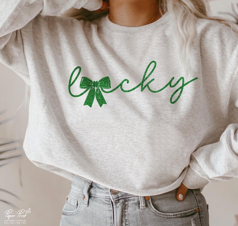 Puede incluir: Una sudadera gris con un dise&ntilde;o de purpurina verde que dice "Lucky" con un lazo verde.