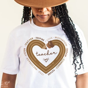 Pu&ograve; includere: Maglietta bianca con un disegno di matita marrone a forma di cuore. La matita &egrave; disegnata con un contorno marrone e ha una gomma marrone. Le parole "teacher" sono scritte al centro del cuore. Le parole "lead, encourage, listen, teacher, inspire, love, motivation, dream, connect, future" sono scritte in cerchio attorno al cuore.