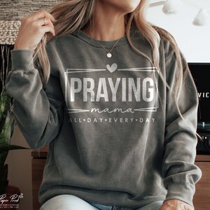 May include: A gray sweatshirt with the text "Praying mama all day every day" in white lettering. The text is inside a white rectangle with a heart at the top.