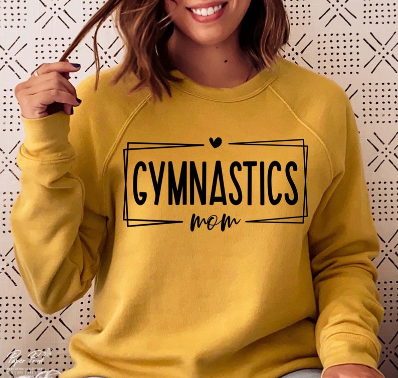 May include: Mustard yellow sweatshirt with the words "GYMNASTICS mom" in black, inside a black rectangular frame. A small black heart is above the word "GYMNASTICS". The sweatshirt has long sleeves and a crew neck.