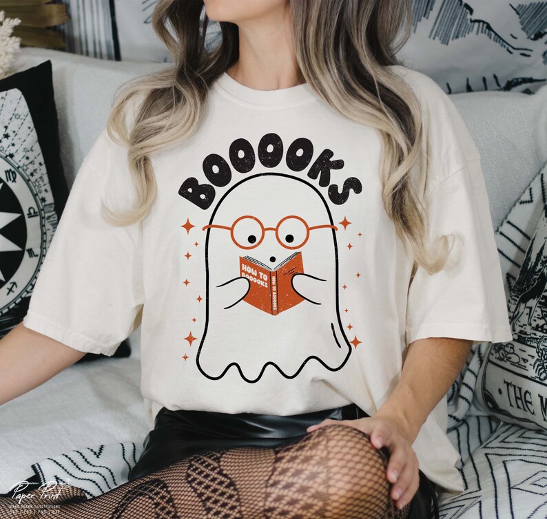 Pode incluir: Camiseta branca com um fantasma usando &oacute;culos e lendo um livro que diz "How to Scare Books". O texto "BOOOKS" est&aacute; acima do fantasma.