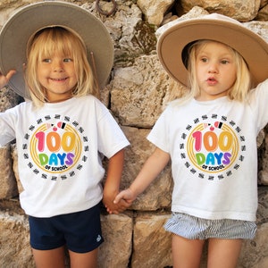May include: Two children wearing white t-shirts with a colorful graphic that says "100 Days of School" and a circle of tally marks around it. The children are wearing straw hats and smiling.