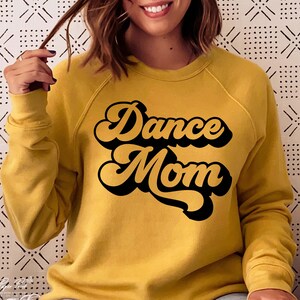 Peut inclure: Un sweat-shirt jaune moutarde avec le texte "Dance Mom" dans une police noire r&eacute;tro.