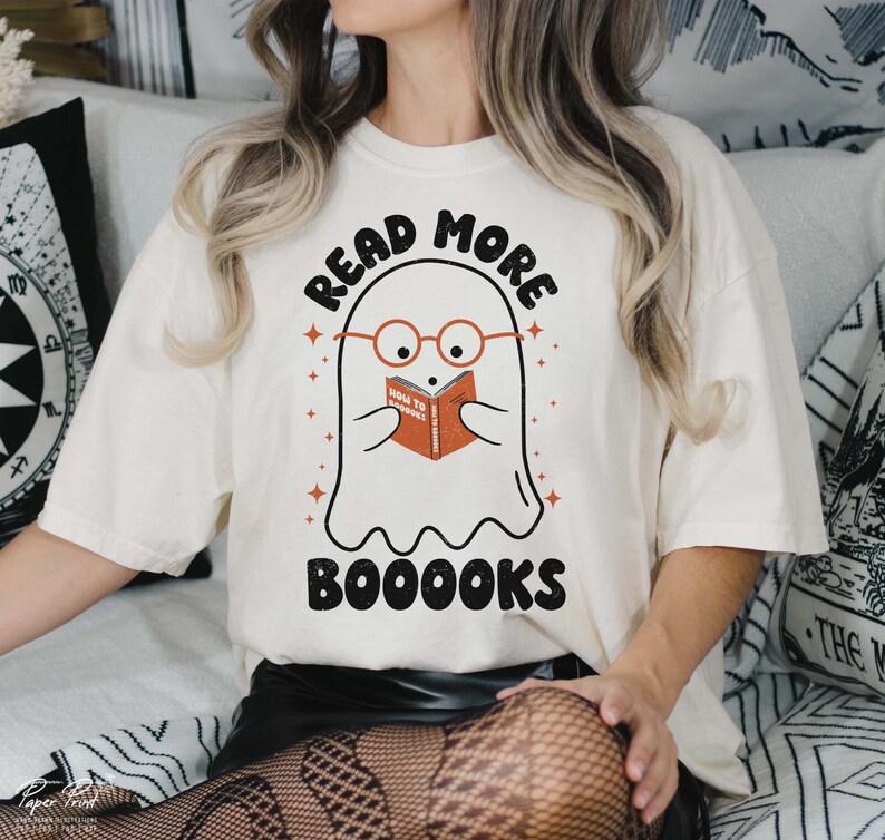 May include: White t-shirt with a ghost wearing glasses and reading a book that says "How to Haunt" with the text "Read More Booooks" above the ghost.