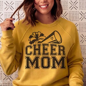 May include: A mustard yellow sweatshirt with a black glitter design of a cheerleader megaphone and pom poms. The text "CHEER MOM" is in black glitter.