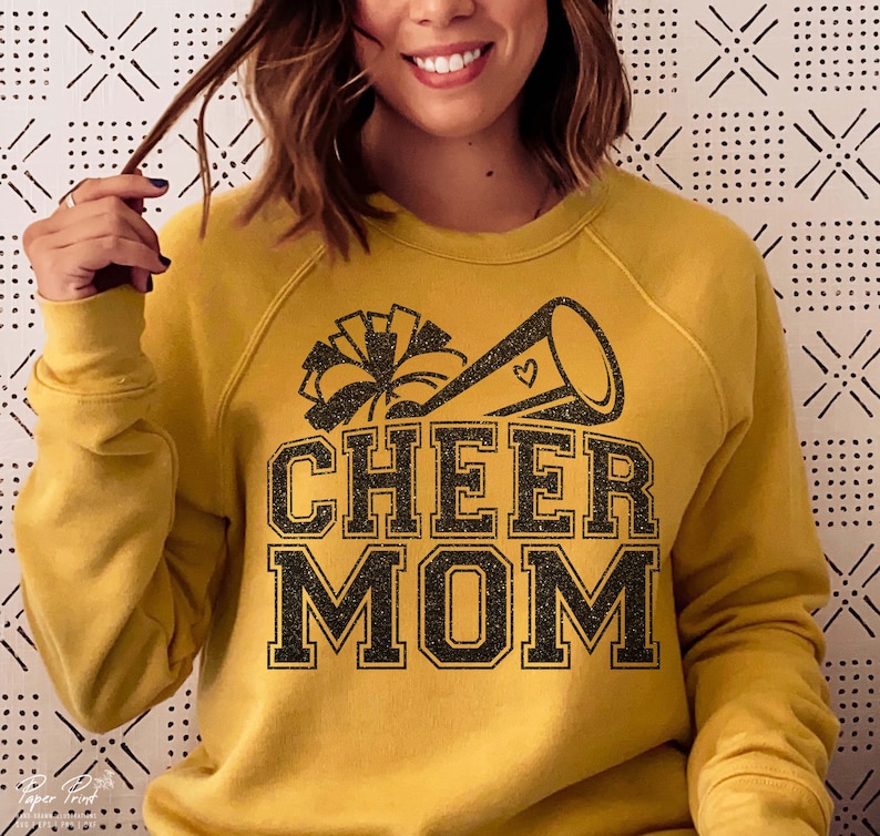 May include: A mustard yellow sweatshirt with a black glitter design of a cheerleader megaphone and pom poms. The text "CHEER MOM" is in black glitter.