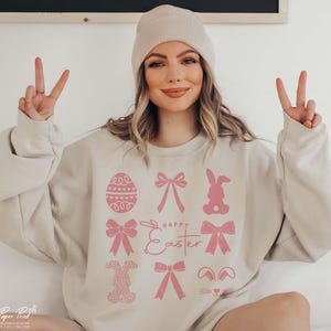 May include: A light beige sweatshirt with a pink graphic design featuring a bunny, eggs, bows, and the text "Happy Easter".