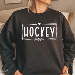 以下が含まれることがあります： 「HOCKEY MOM」という白い文字が書かれた黒いスウェットシャツ。HOCKEYの上には小さなハートマークがあります。