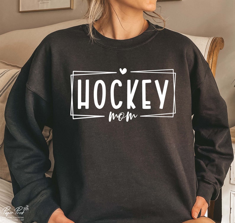 以下が含まれることがあります： 「HOCKEY MOM」という白い文字が書かれた黒いスウェットシャツ。HOCKEYの上には小さなハートマークがあります。