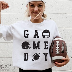 May include: White sweatshirt with black text that reads "GAME DAY" with football helmet and football graphics. A person is holding a brown football.