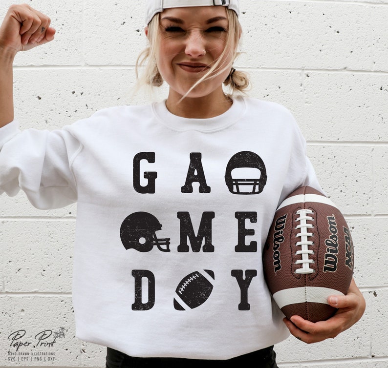 May include: White sweatshirt with black text that reads "GAME DAY" with football helmet and football graphics. A person is holding a brown football.