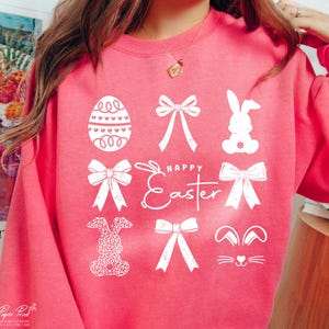 May include: Pink sweatshirt with a white graphic design featuring Easter-themed elements like bunnies, bows, and eggs. The text "Happy Easter" is also included in the design.