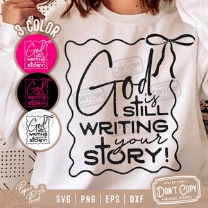 May include: A white sweatshirt with a black glitter design that says "God is still writing your story!" in a bold, cursive font. The design is surrounded by a black border with a decorative bow at the top. The sweatshirt is a blank canvas for a custom design.