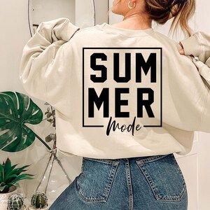 May include: A beige sweatshirt with the words "SUMMER Mode" printed in black within a black square. The sweatshirt is paired with blue denim jeans. The image is taken from the back, showing the sweatshirt's design.