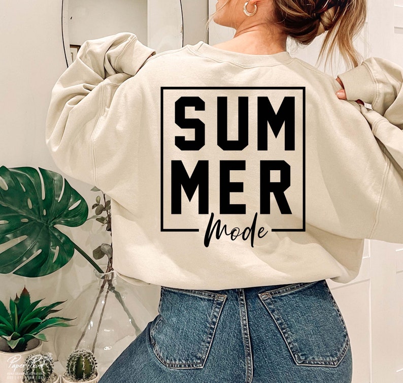 May include: A beige sweatshirt with the words "SUMMER Mode" printed in black within a black square. The sweatshirt is paired with blue denim jeans. The image is taken from the back, showing the sweatshirt's design.