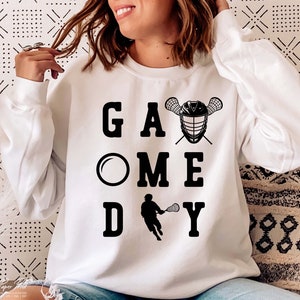 May include: White sweatshirt with black text that reads "Game Day" with a lacrosse stick and helmet graphic.