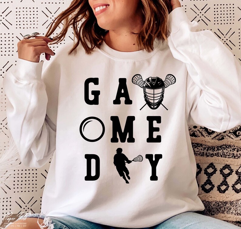 May include: White sweatshirt with black text that reads "Game Day" with a lacrosse stick and helmet graphic.
