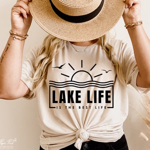 May include: Beige t-shirt with a black graphic of a sun over water, two birds, and the text "LAKE LIFE IS THE BEST LIFE". The shirt is tied at the waist and worn with a straw hat and bracelets.