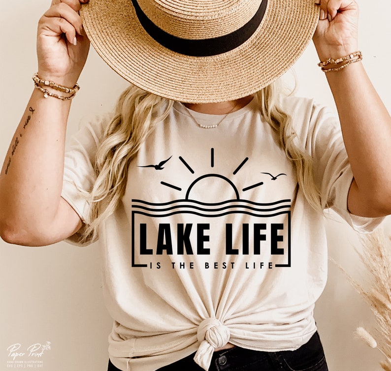 May include: Beige t-shirt with a black graphic of a sun over water, two birds, and the text "LAKE LIFE IS THE BEST LIFE". The shirt is tied at the waist and worn with a straw hat and bracelets.