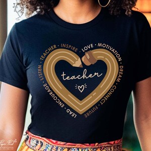 Pu&ograve; includere: Una maglietta nera con un design a forma di cuore dorato che presenta una matita e la parola "teacher" all'interno del cuore. Il cuore &egrave; circondato dalle parole "Teacher, Inspire, Love, Motivation, Dream, Connect, Future, Lead, Encourage, Listen".