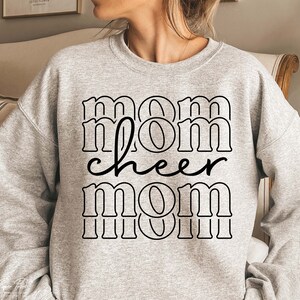May include: A light grey sweatshirt featuring the text "mom cheer mom" in a black, bold, outlined font. The word "cheer" is in a cursive font. The sweatshirt has a crew neck.