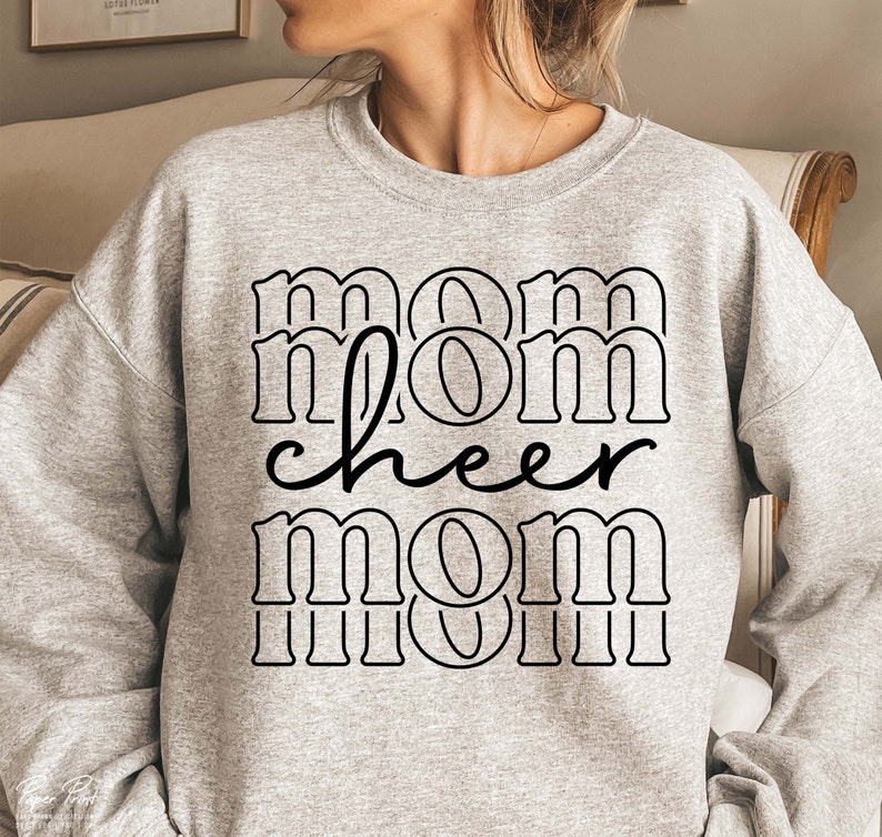 May include: A light grey sweatshirt featuring the text "mom cheer mom" in a black, bold, outlined font. The word "cheer" is in a cursive font. The sweatshirt has a crew neck.