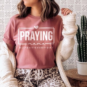 May include: A pink t-shirt with the text "Praying mama all day every day" in white lettering. The text is framed in a white rectangle with a small heart above it.