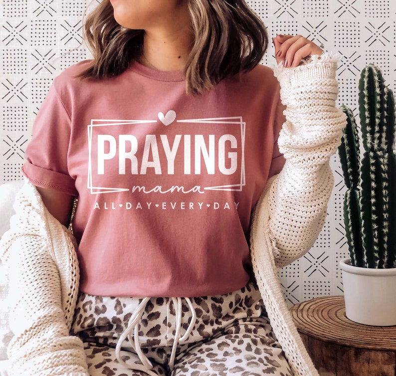 May include: A pink t-shirt with the text "Praying mama all day every day" in white lettering. The text is framed in a white rectangle with a small heart above it.