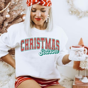 May include: White sweatshirt with red and green text that reads "Christmas Season". The sweatshirt is being worn by a person holding a white ceramic mug with a Santa Claus face.
