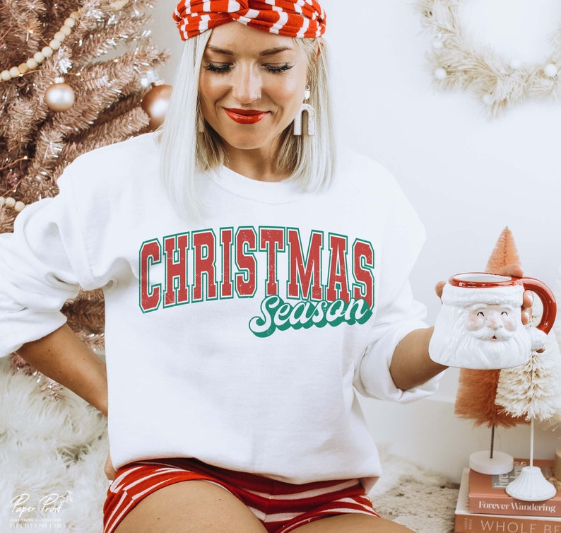 May include: White sweatshirt with red and green text that reads "Christmas Season". The sweatshirt is being worn by a person holding a white ceramic mug with a Santa Claus face.