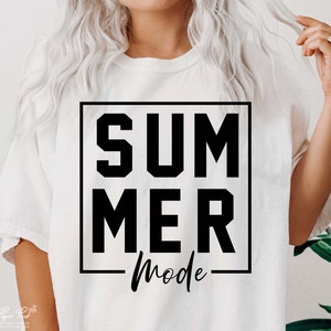 May include: A white t-shirt with the words "SUMMER MODE" in a black, bold, block font inside a black square. The word "Mode" is in a cursive font. The shirt is being worn by a person.