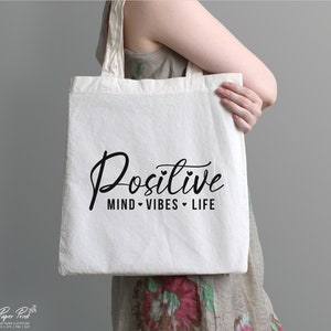 May include: A white canvas tote bag with black text that reads "Positive Mind Vibes Life".