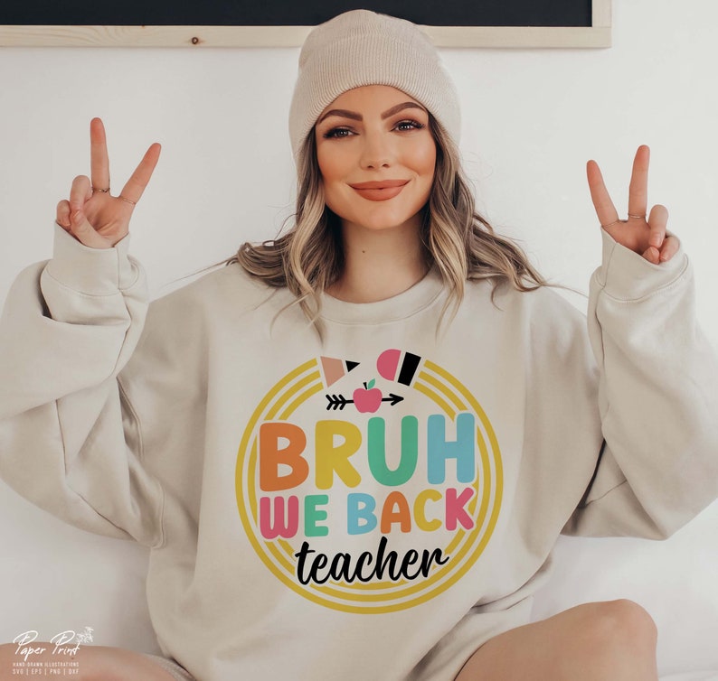 Puede incluir: Una sudadera de color beige claro con un dise&ntilde;o gr&aacute;fico colorido que dice "BRUH WE BACK teacher" en un c&iacute;rculo con un borde de arco&iacute;ris.