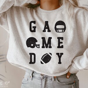 May include: A gray sweatshirt with the text "GAME DAY" in black block letters. The text is surrounded by images of football helmets and a football.