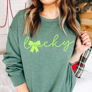 Puede incluir: Una sudadera verde con la palabra "lucky" escrita en letra cursiva con un lazo verde en el medio de la palabra.