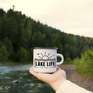 May include: White enamel mug with a black rim and handle, featuring the text "LAKE LIFE IS THE BEST LIFE" in a black rectangular design with a sun and birds. The mug is held in front of a blurred natural background.