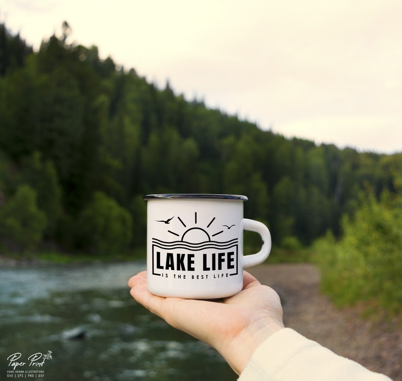 May include: White enamel mug with a black rim and handle, featuring the text "LAKE LIFE IS THE BEST LIFE" in a black rectangular design with a sun and birds. The mug is held in front of a blurred natural background.