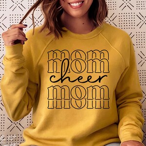 May include: Mustard yellow sweatshirt with the words "mom cheer mom" in black lettering. The text is stacked with a retro font. The sweatshirt has long sleeves and a crew neck.