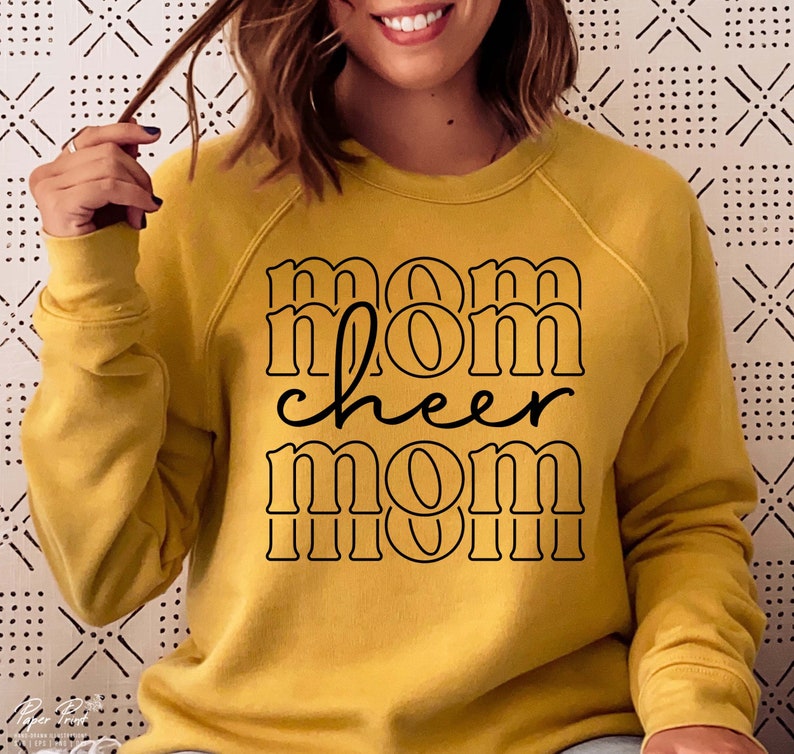 May include: Mustard yellow sweatshirt with the words "mom cheer mom" in black lettering. The text is stacked with a retro font. The sweatshirt has long sleeves and a crew neck.