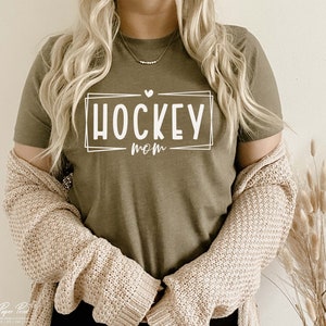 以下が含まれることがあります： 白い枠の中に「HOCKEY MOM」という文字が書かれた緑色のTシャツです。
