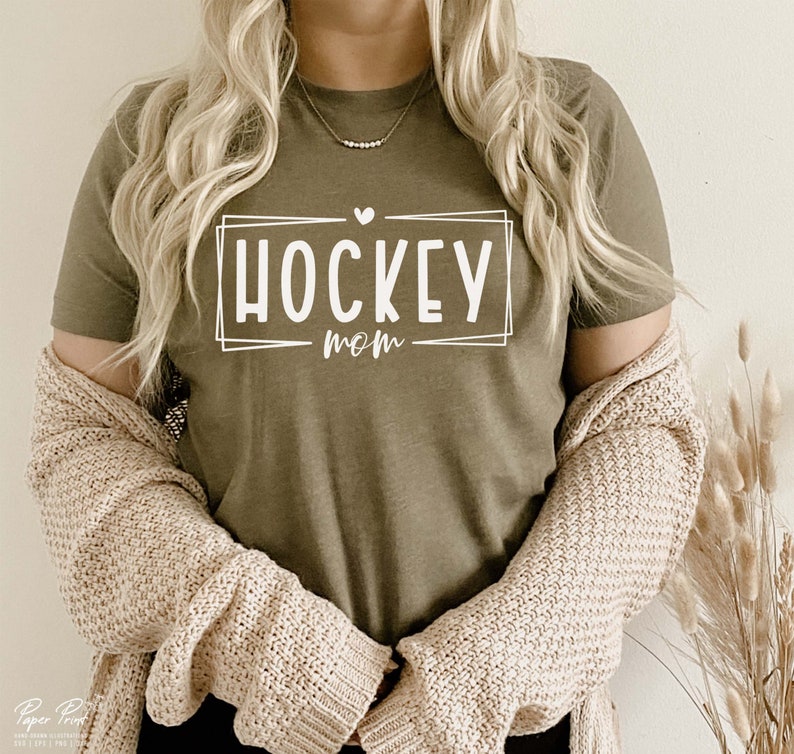 以下が含まれることがあります： 白い枠の中に「HOCKEY MOM」という文字が書かれた緑色のTシャツです。