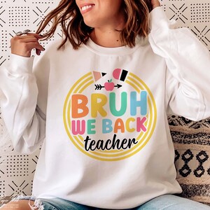 Puede incluir: Sudadera blanca con un gr&aacute;fico colorido que dice "Bruh We Back Teacher" con una flecha arco&iacute;ris y una manzana.