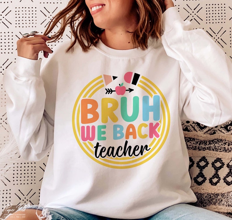 Puede incluir: Sudadera blanca con un gr&aacute;fico colorido que dice "Bruh We Back Teacher" con una flecha arco&iacute;ris y una manzana.