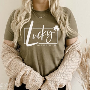 May include: A green t-shirt with white text that says "Lucky" with a shamrock and "All Day Every Day" underneath. The shirt is being worn by a person with long blonde hair.