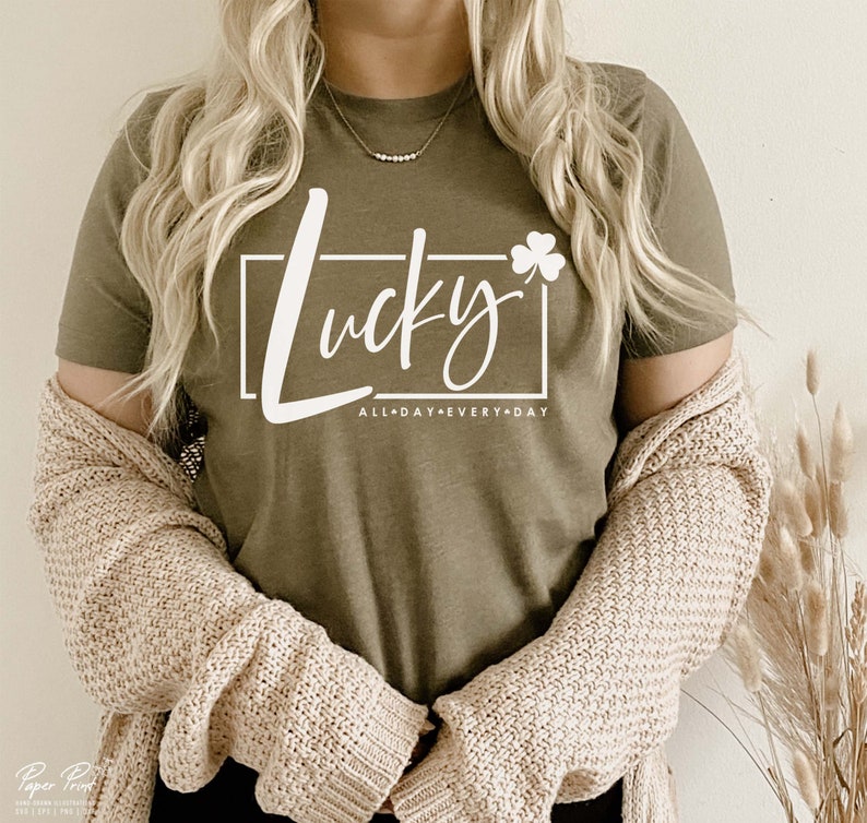 May include: A green t-shirt with white text that says "Lucky" with a shamrock and "All Day Every Day" underneath. The shirt is being worn by a person with long blonde hair.